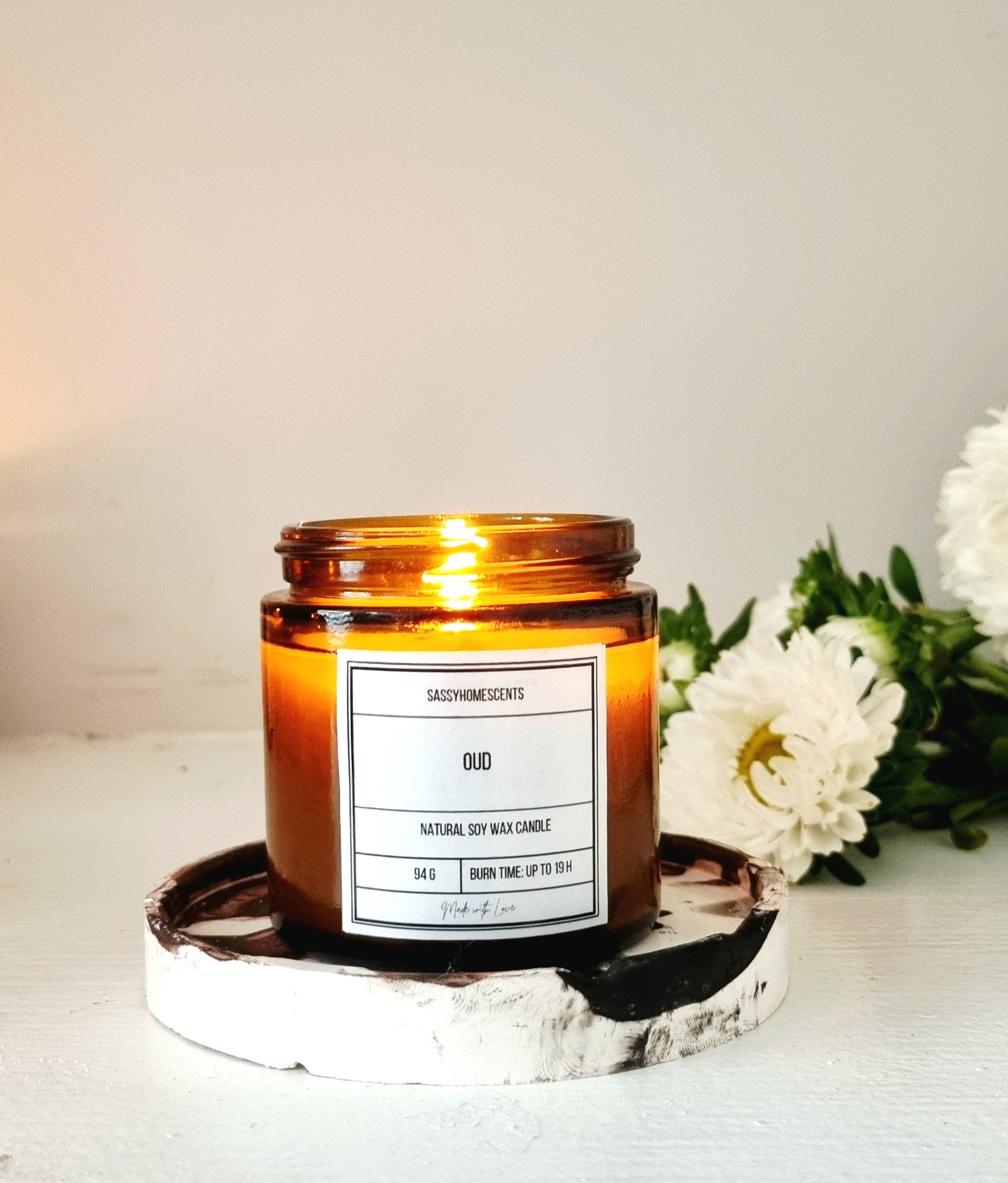 Amber candle with a label on a white surface with flowers in the background
